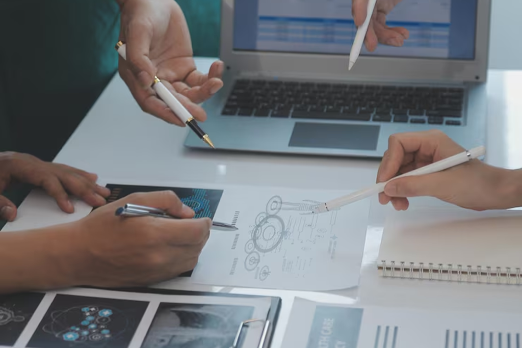 Team collaborating over research documents and technical diagrams with a laptop, representing NLP consultant services for summarizing medical research papers and extracting key findings to support faster healthcare decision-making.