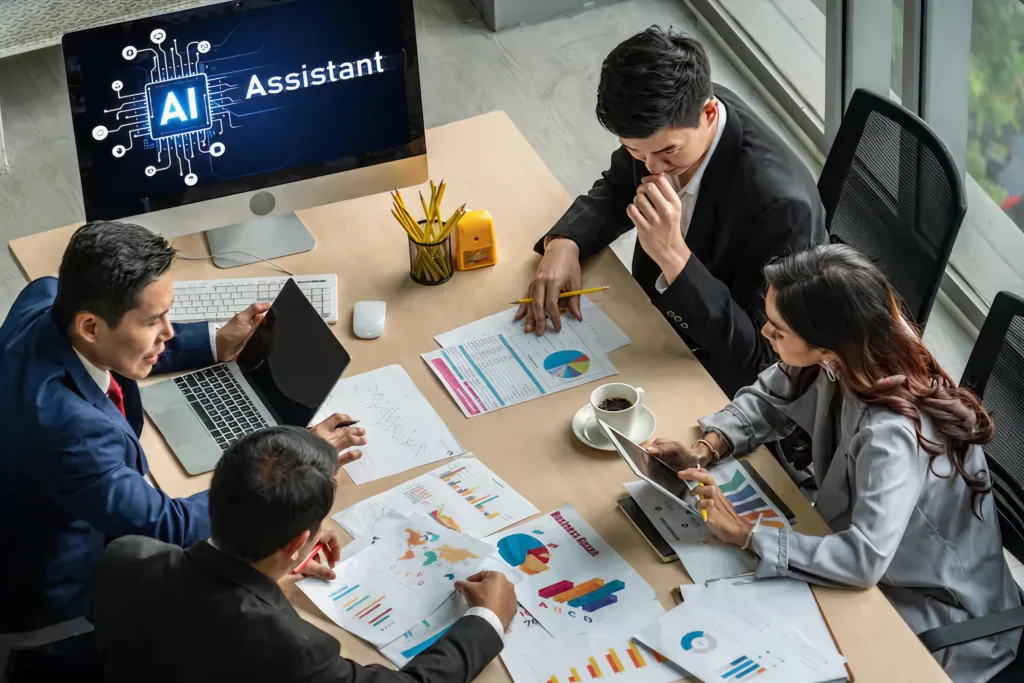 Business team analyzing charts and discussing AI strategy around a desk, with a screen displaying “AI Assistant,” representing AI outsourcing services that support end-to-end solution design, development, and scalability.