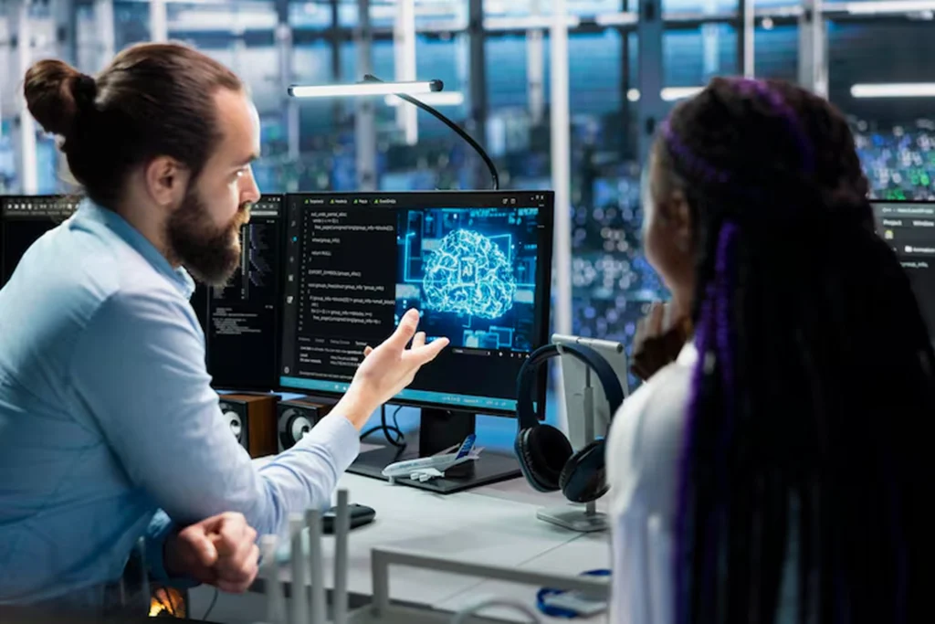 AI developers discussing system performance in a modern office, with AI code and neural graphics on the screen, highlighting continuous monitoring, model retraining, and long-term optimization offered by AI outsourcing services.