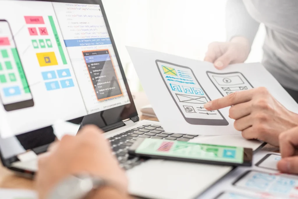Two people discussing and pointing at wireframes for a mobile app design, while the laptop screen displays code and UI elements, showcasing the process of UI component and design implementation.